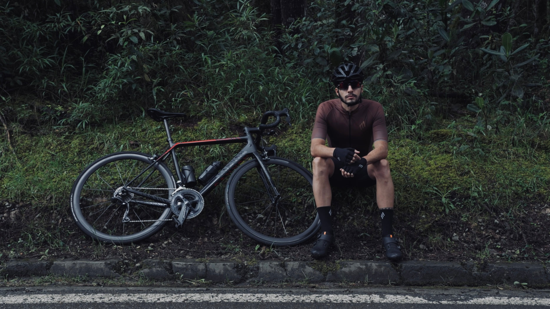 Foto de ciclista de ruta sentado al lado de la ruta. LLeva puesto ropas de ciclismo de marcas colombianas.